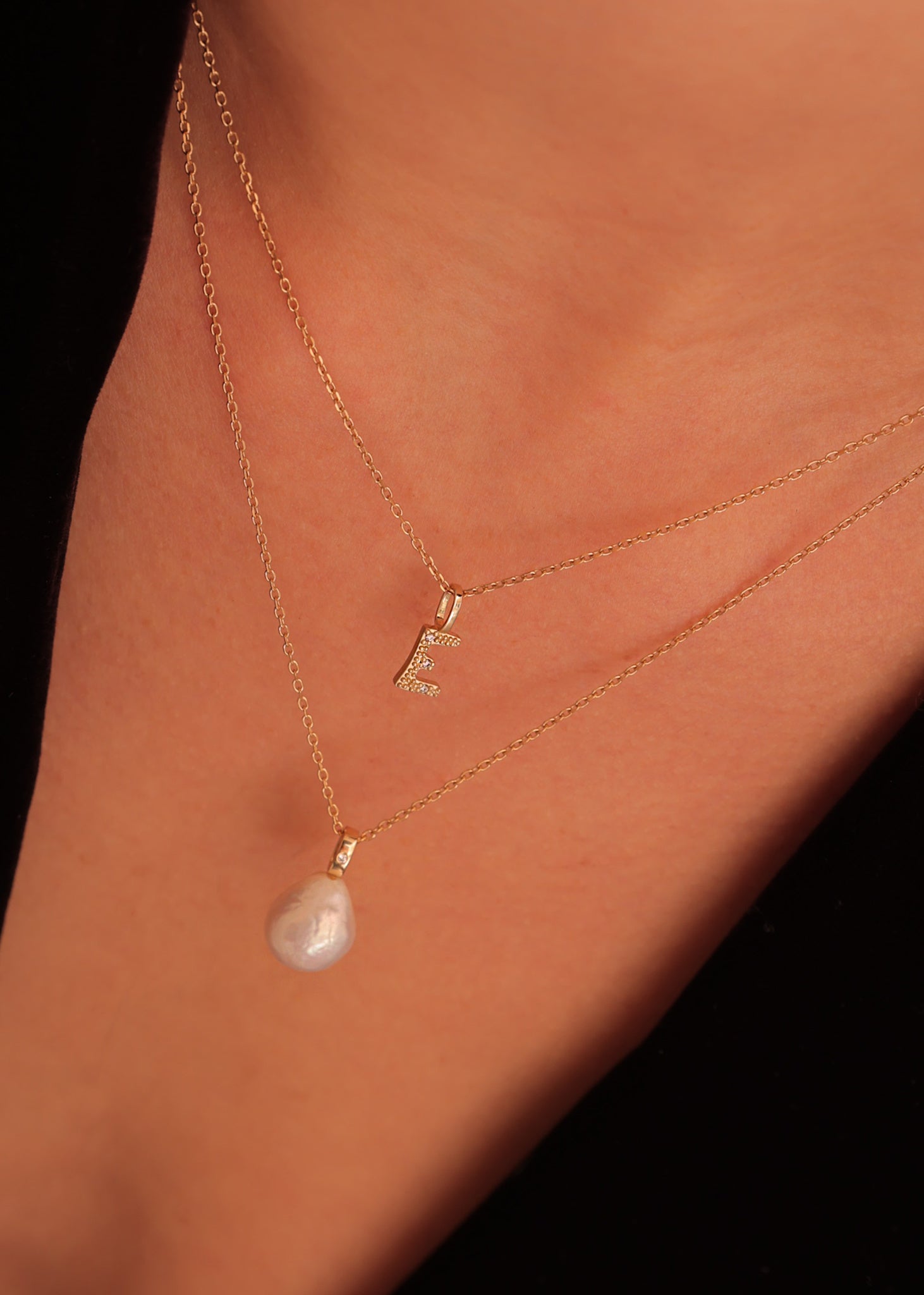 Kvinde med to vedhæng til halskæder i ægte guld. Det ene vedhæng er et bogstavsvedhæng med bogstavet E sat med tre diamanter. Det andet vedhæng er et perlevedhæng til halskæder med en diamant i øksenet i guld